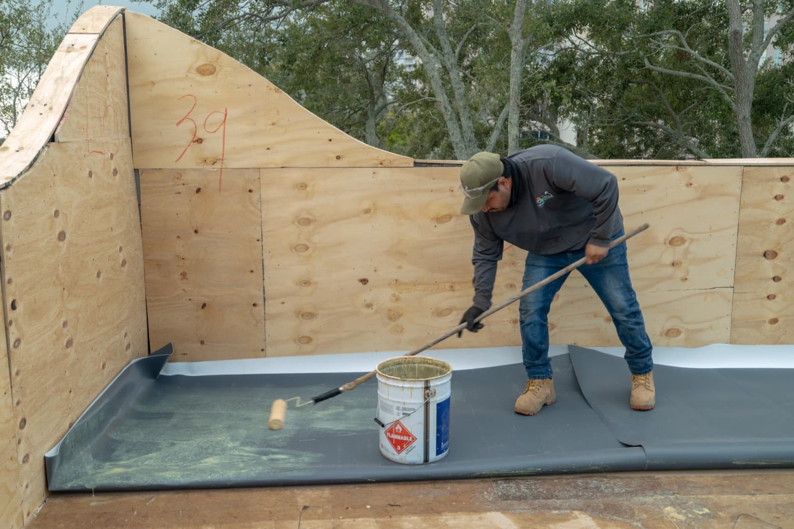 A roofer in a ball cap and work boots using a long-handled roller to spread coating over a black low-slope roof membrane, with a paint bucket at his feet and plywood parapet walls around him.