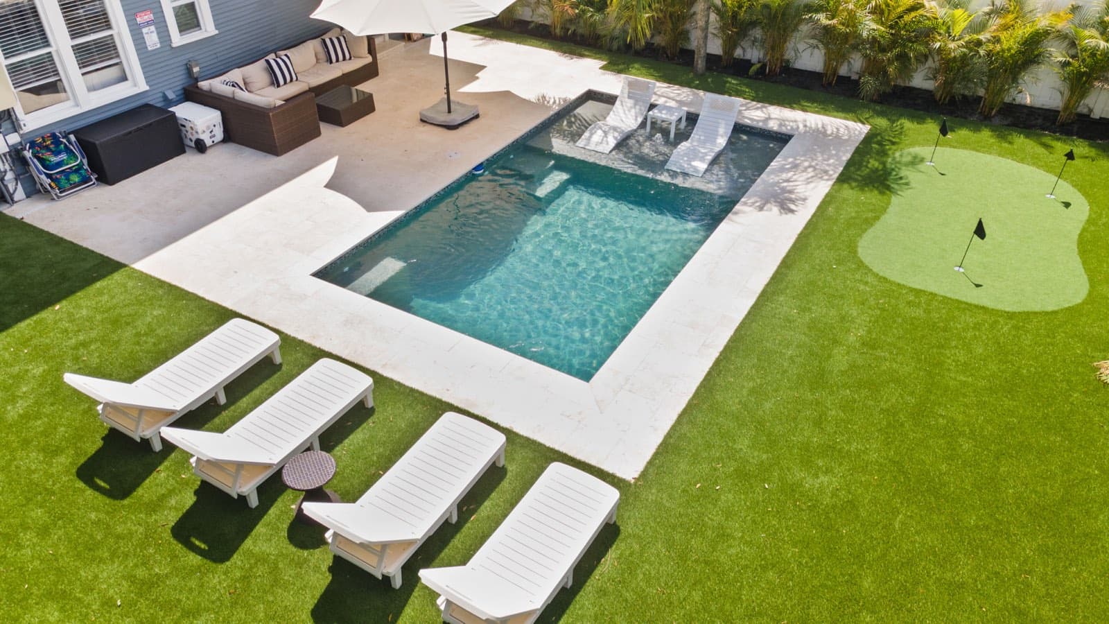 Aerial view of a finished rectangular pool with a tanning ledge, white pavers, lounge chairs, and a putting green in a Florida backyard.