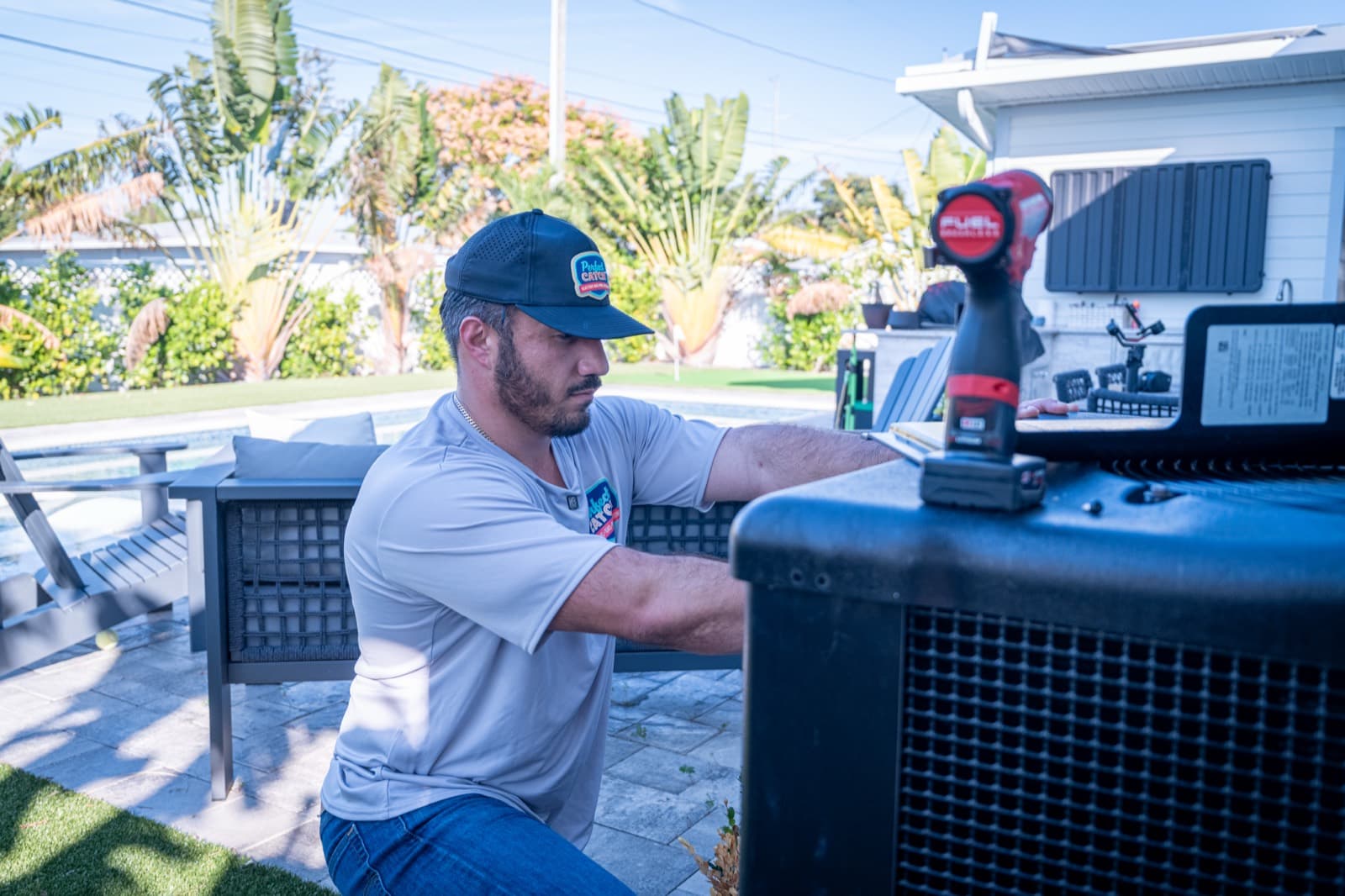 An HVAC technician in a company ball cap crouched beside a black outdoor heat pump unit in a Tampa Bay backyard, with a pool and palm trees behind him.