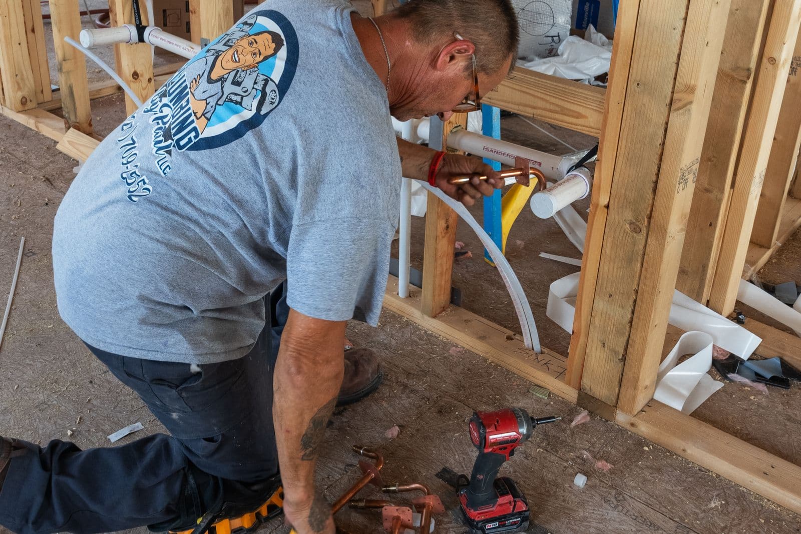 A plumber in a grey company t-shirt kneels inside a framed wall, fitting copper and white PEX lines, with a red cordless drill and copper fittings on the plywood floor beside him.