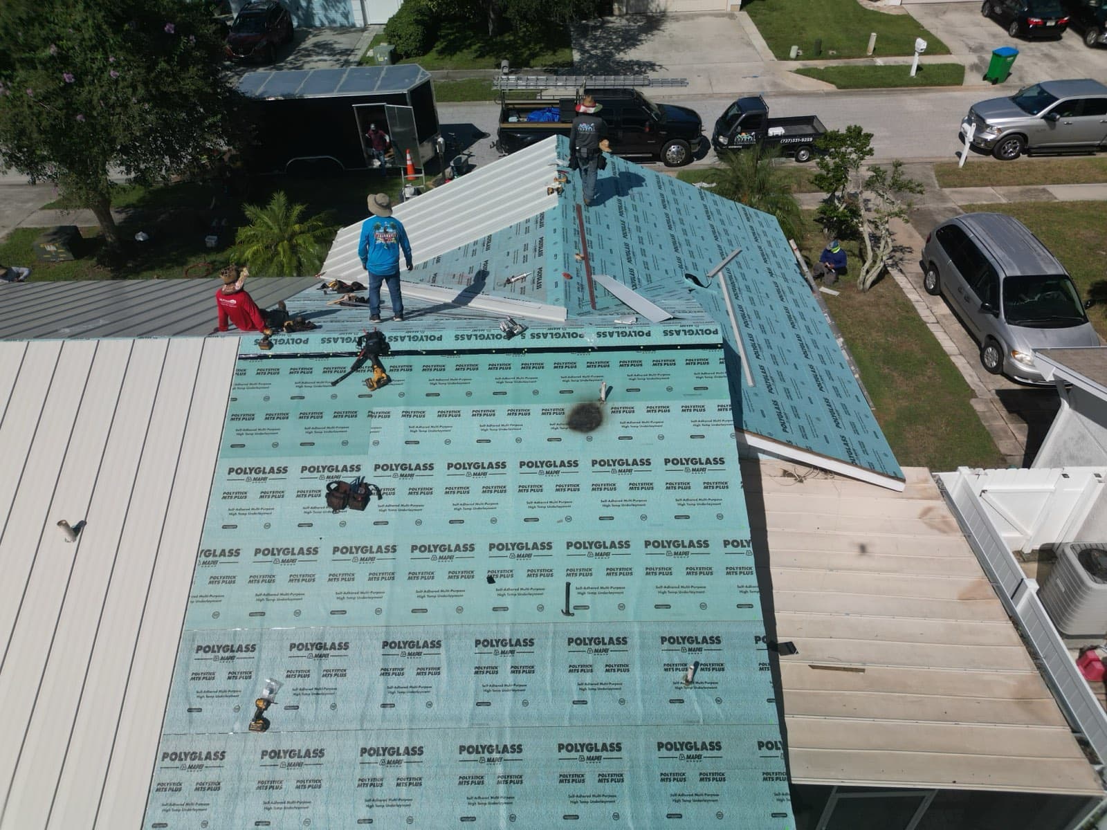 Drone view of a roofing crew on a residential re-roof, synthetic underlayment down on one slope and new metal panels going on the other