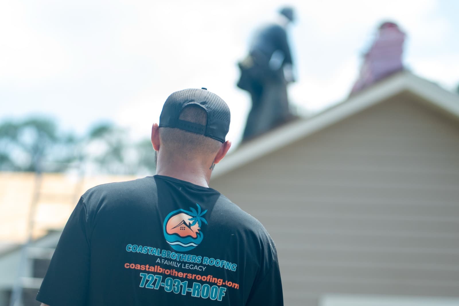 A roofer in a black Coastal Brothers Roofing shirt and cap, seen from behind, watches a crew work on roofs with exposed decking in the background.