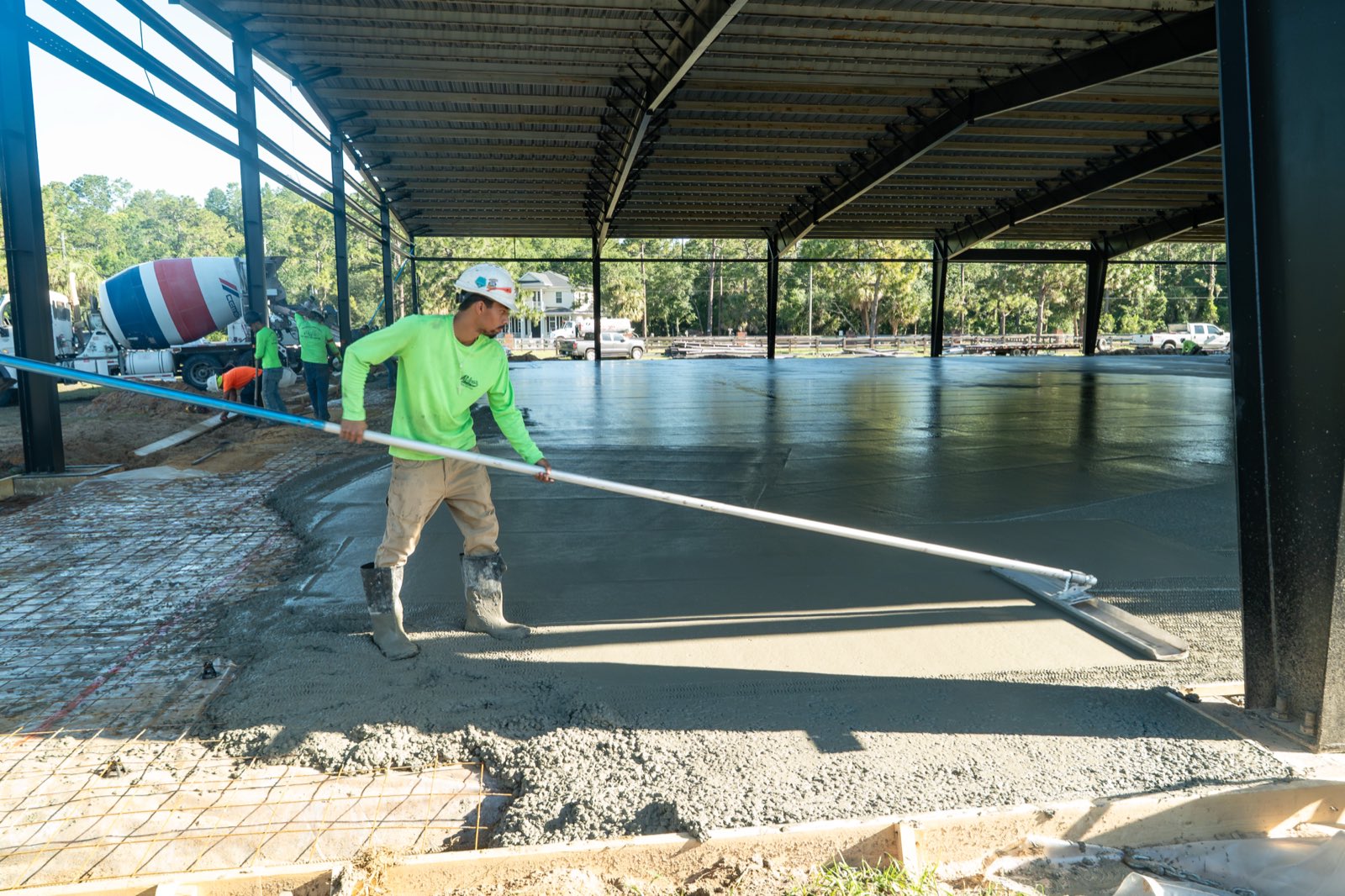 A concrete finisher in a green long-sleeve shirt pulling a screed across a wet slab under a steel-framed structure, with a mixer truck parked behind him.