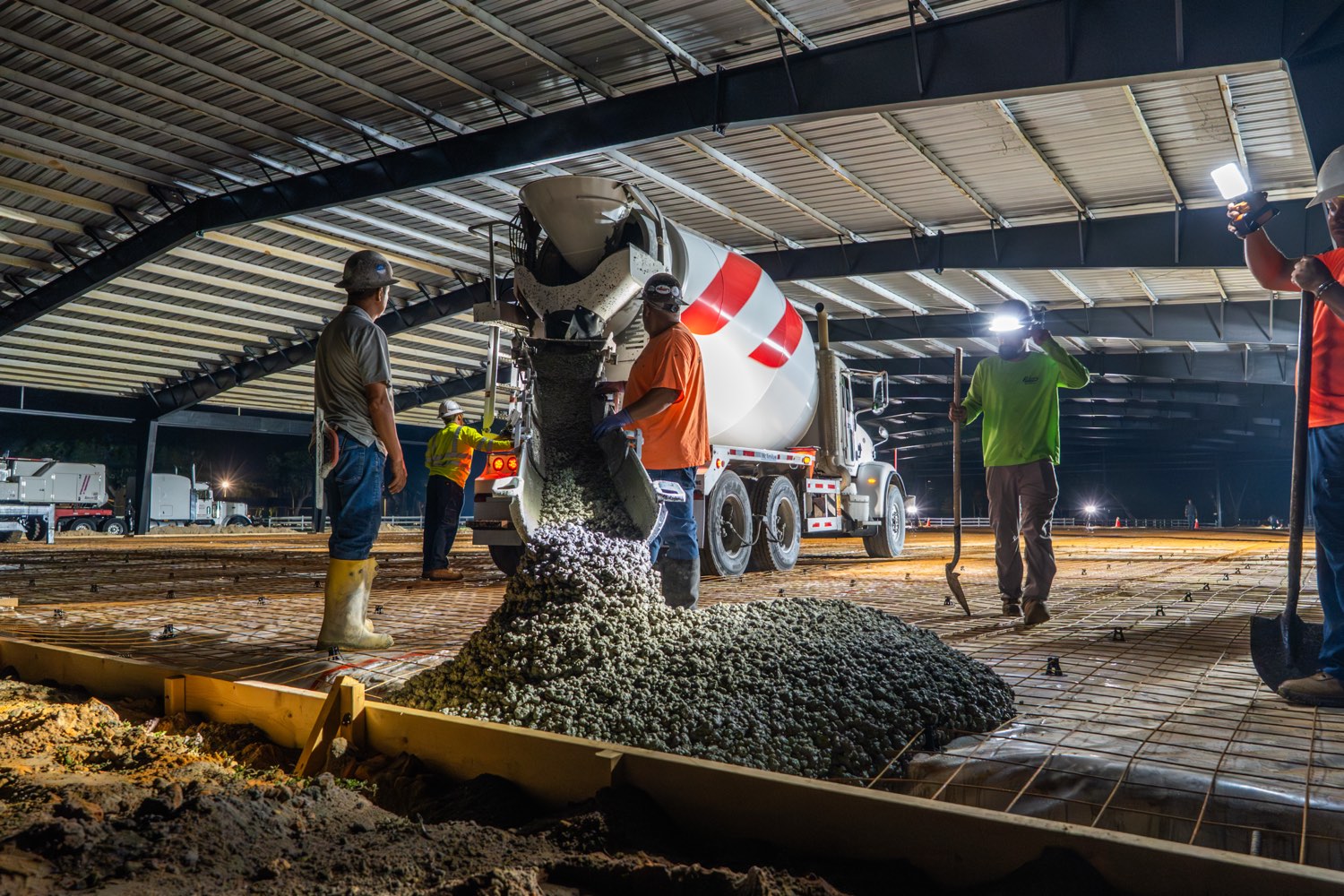 Crew placing concrete from a mixer truck under a steel structure at night, lit by work lights