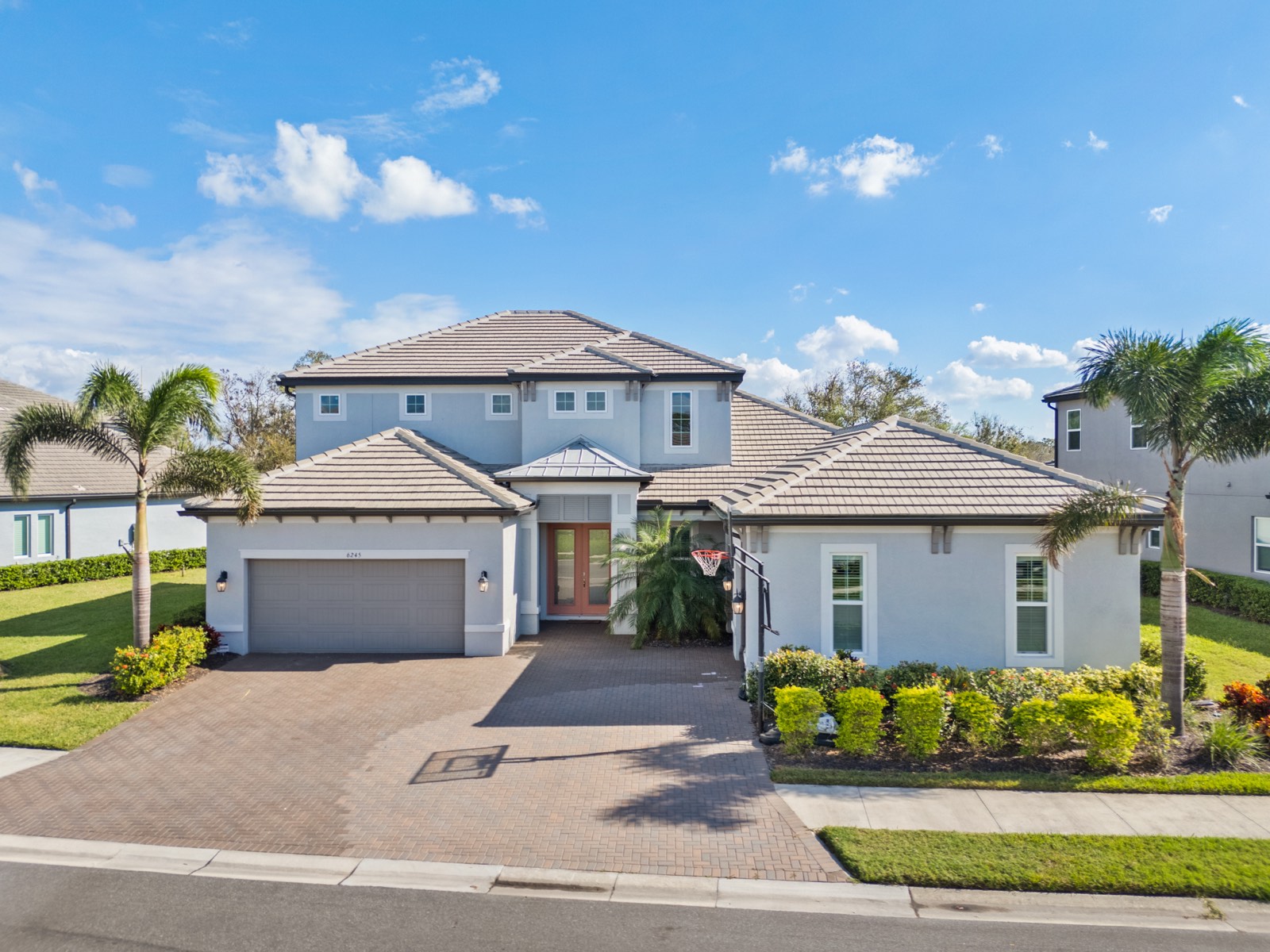 Elevated drone view of a two-story tile-roof home with paver driveway and palms on a sunny day