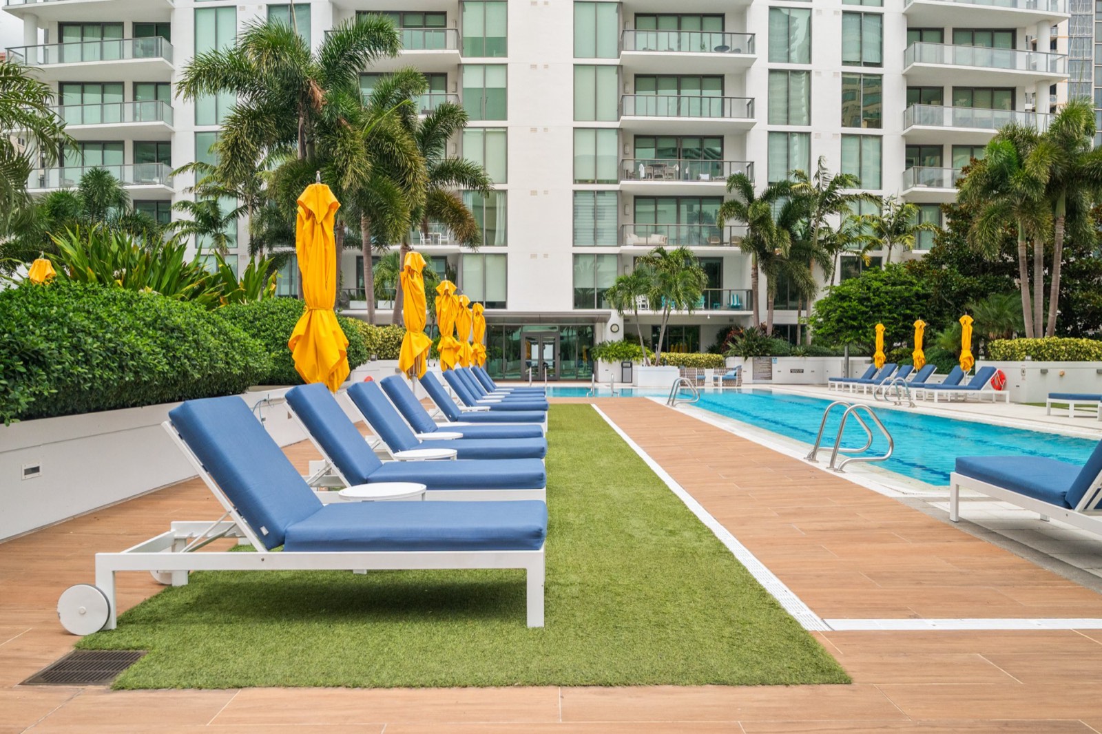 Resort-style condo pool deck with blue loungers, yellow umbrellas and palms against the building facade